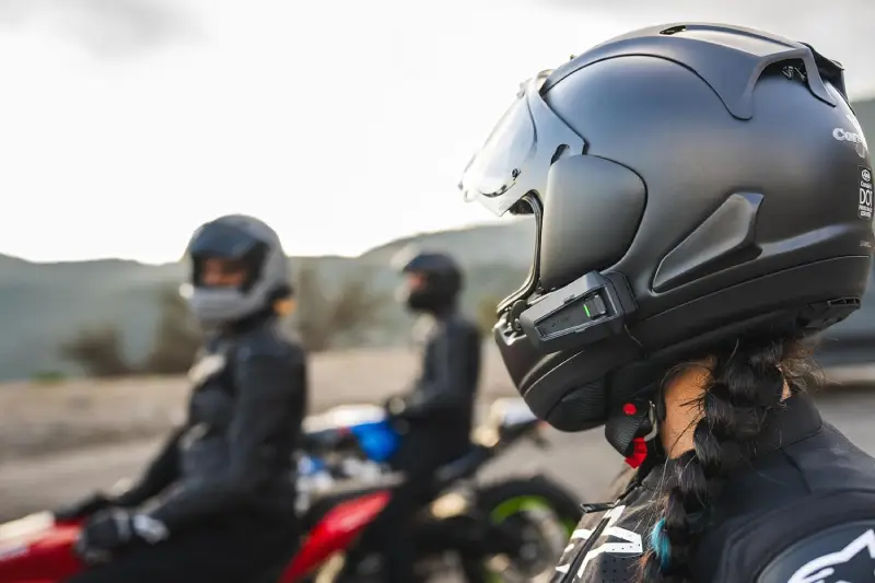 A close-up rear-quarter view of a motorcyclist wearing a matte black helmet equipped with a cardo packtalk pro communication system, with two other riders on motorcycles visible in the blurred background.